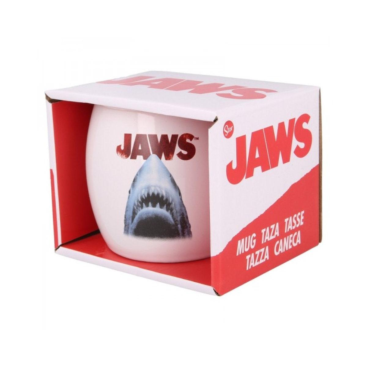 Una taza de Tiburón en una caja. La taza tiene una imagen de tiburón y el texto 'JAWS'. La caja es blanca y roja con la marca 'JAWS'.