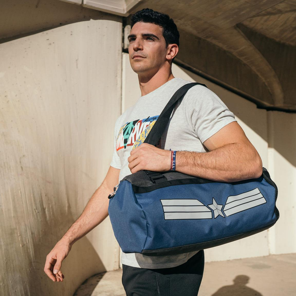 Hombre caminando, llevando una bolsa de gimnasio azul con rayas blancas. Viste una camiseta de Marvel y una pulsera. El fondo es una estructura de concreto.