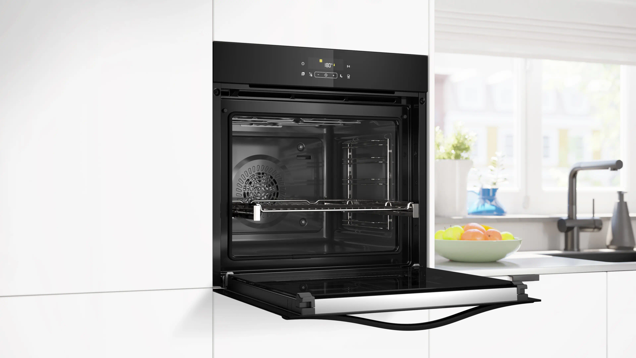 Horno negro con puerta abierta, interior visible. Ambiente de cocina con ventana, frutero y grifo al fondo.