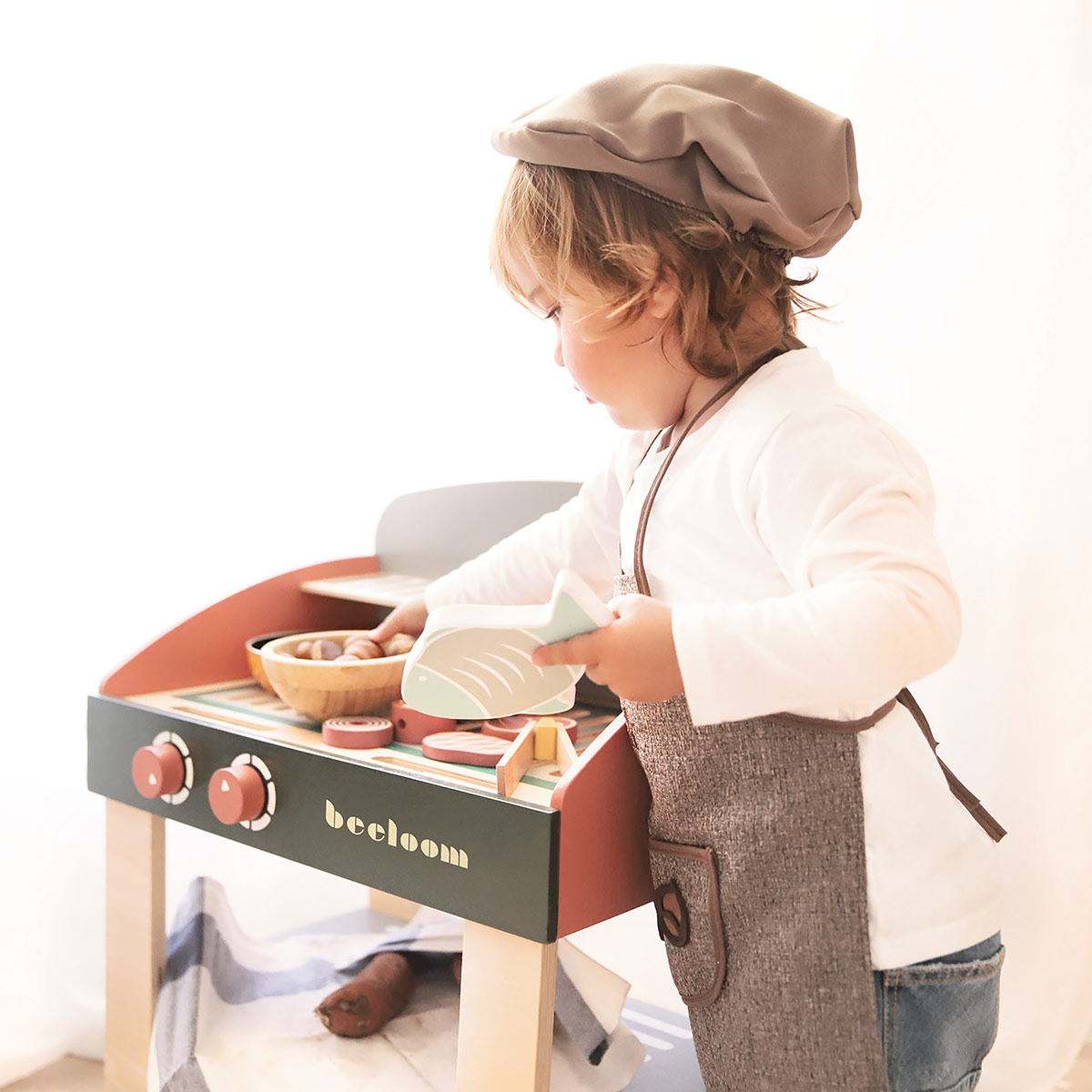Un niño con gorro y delantal de chef juega con una cocina de juguete. El juguete tiene comida de madera y el niño sostiene un pez.