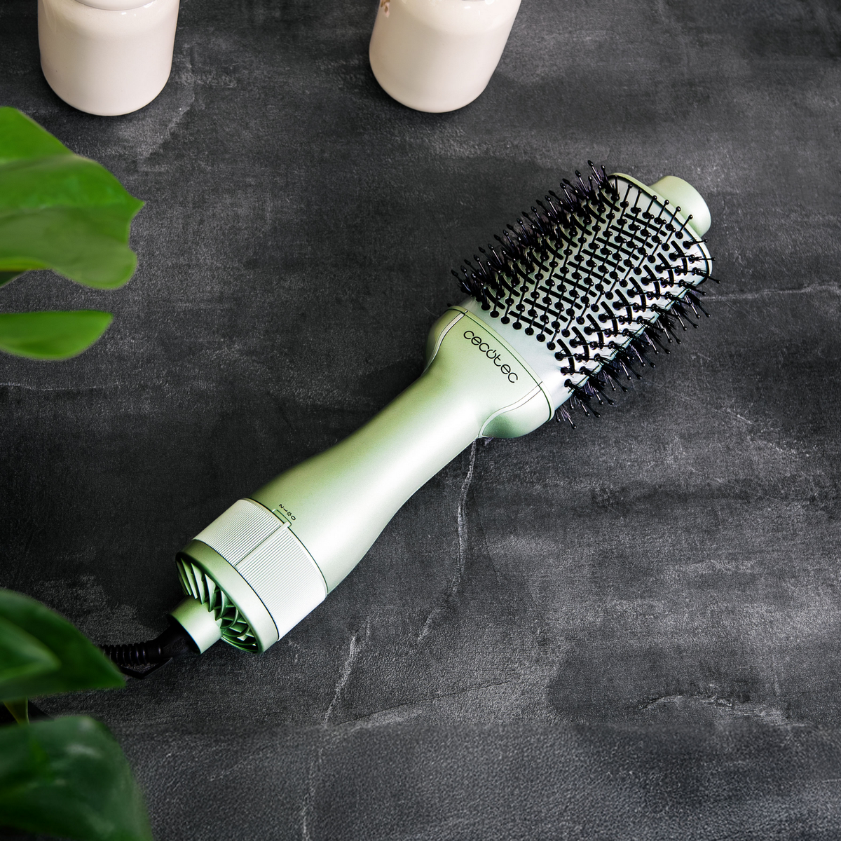 Un asciugacapelli verde con setole nere è su una superficie grigio scuro, accanto a due barattoli bianchi e foglie verdi.
