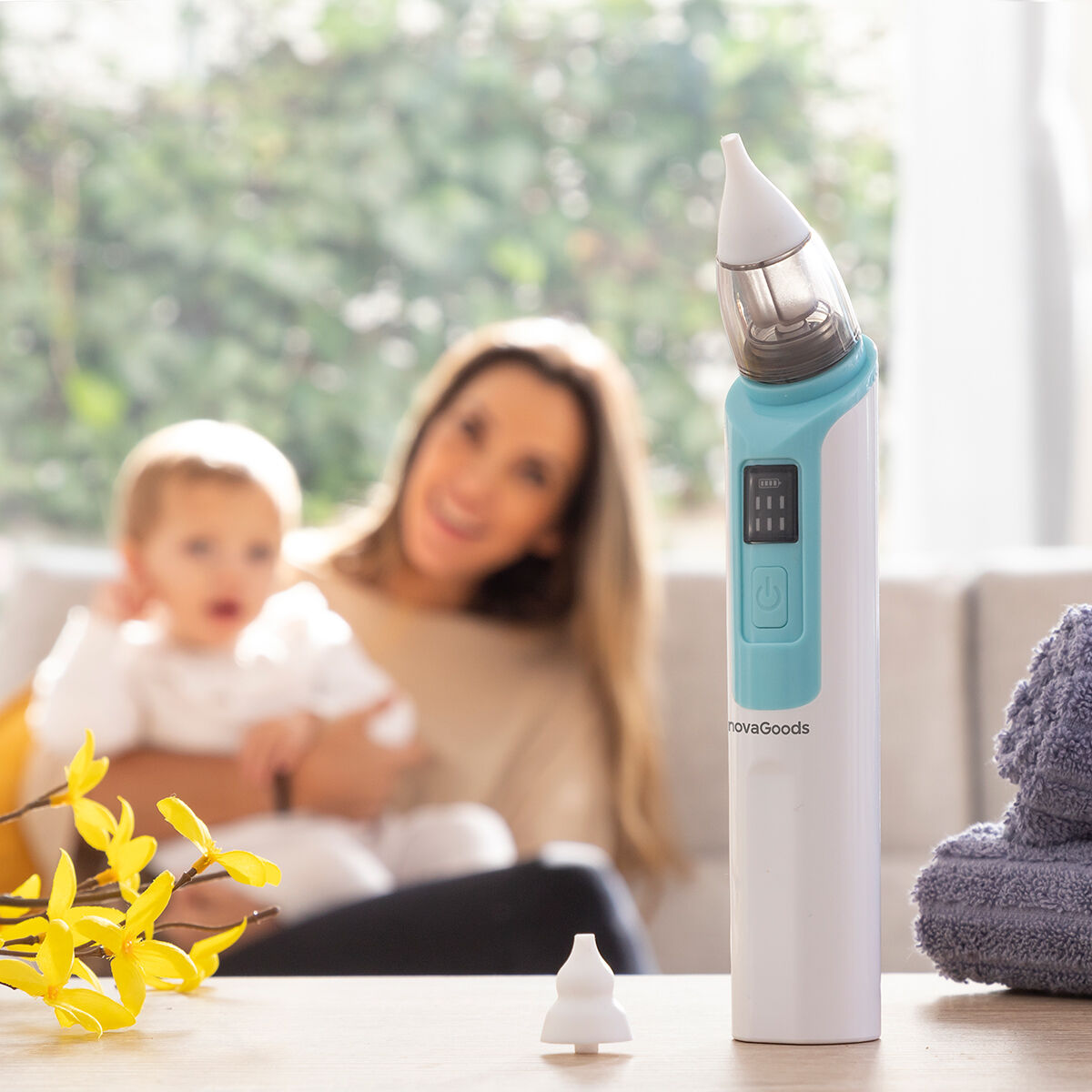 Un aspirador nasal para bebés sobre una mesa, con una madre y un niño borrosos en el fondo. Flores amarillas en primer plano y una toalla a la derecha.