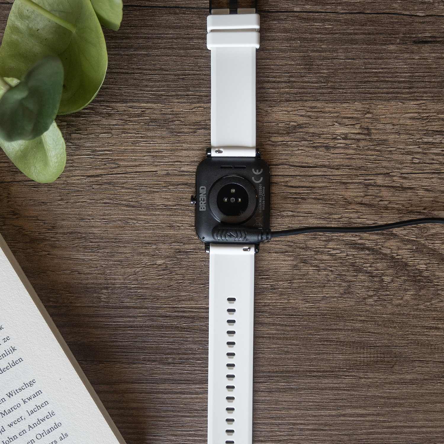 Een witte smartwatch ligt op een houten oppervlak met een blad en een boek. Het horloge wordt opgeladen. De achtergrond is een houten tafel.
