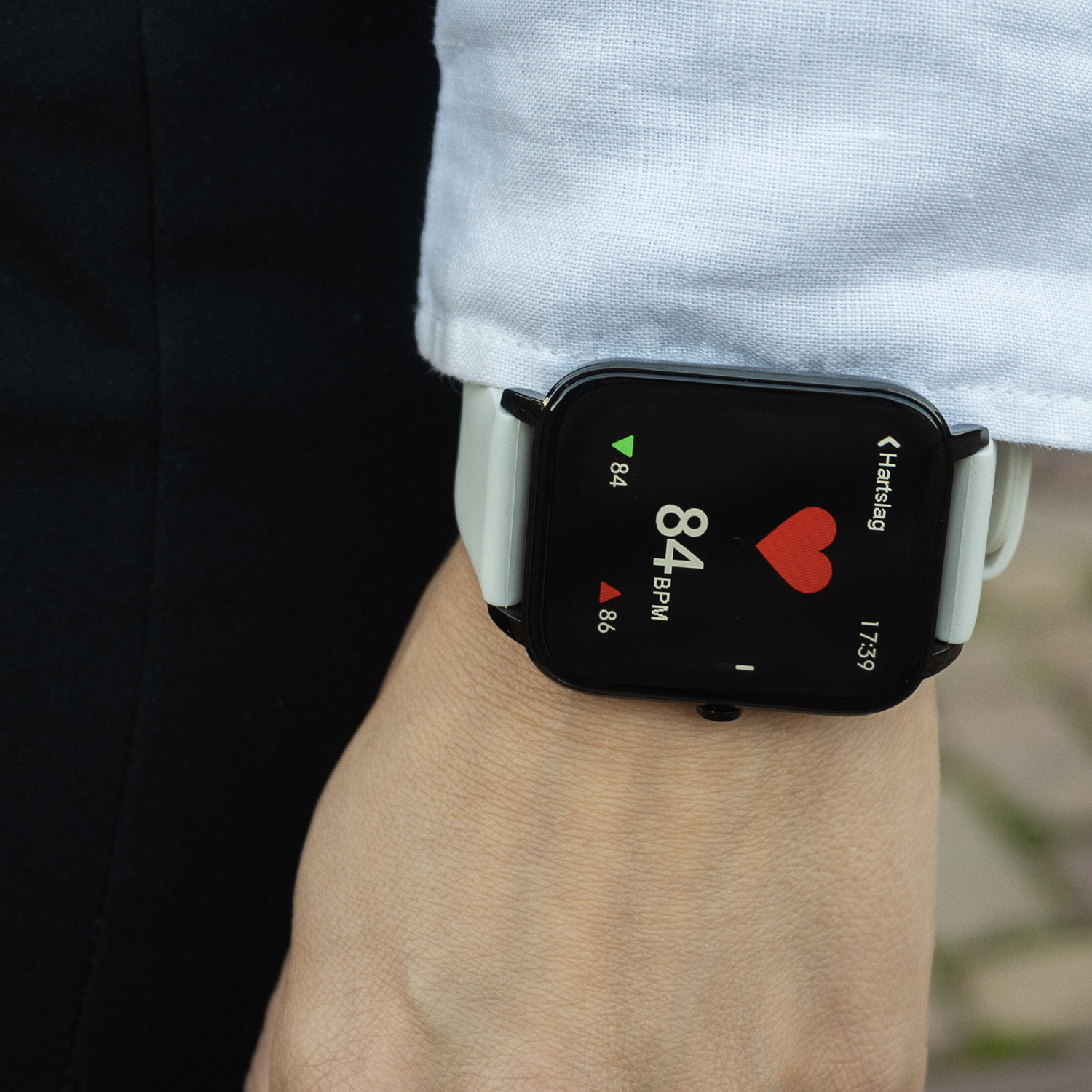 Een pols met een smartwatch die de hartslag weergeeft, 84 BPM. Het horloge heeft een zwart scherm, witte band, op een pols.