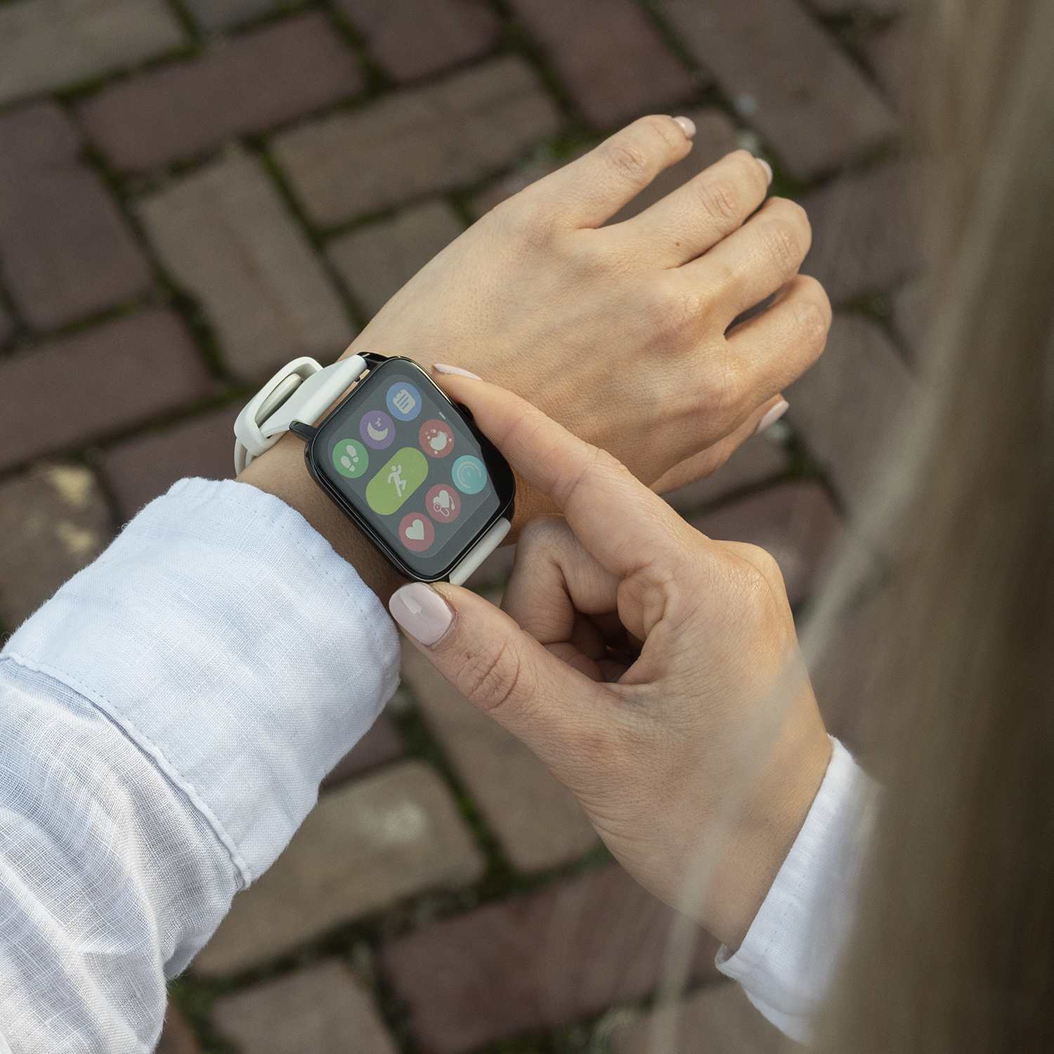 Een vrouw draagt een witte smartwatch. Ze raakt het scherm aan met haar rechterhand. De achtergrond is een bakstenen pad.