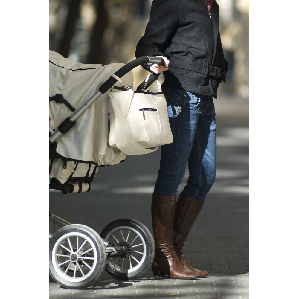 Frau mit Kinderwagen und cremefarbener Tasche. Sie trägt Jeans, eine schwarze Jacke und braune Stiefel. Der Kinderwagen hat weiße Räder.