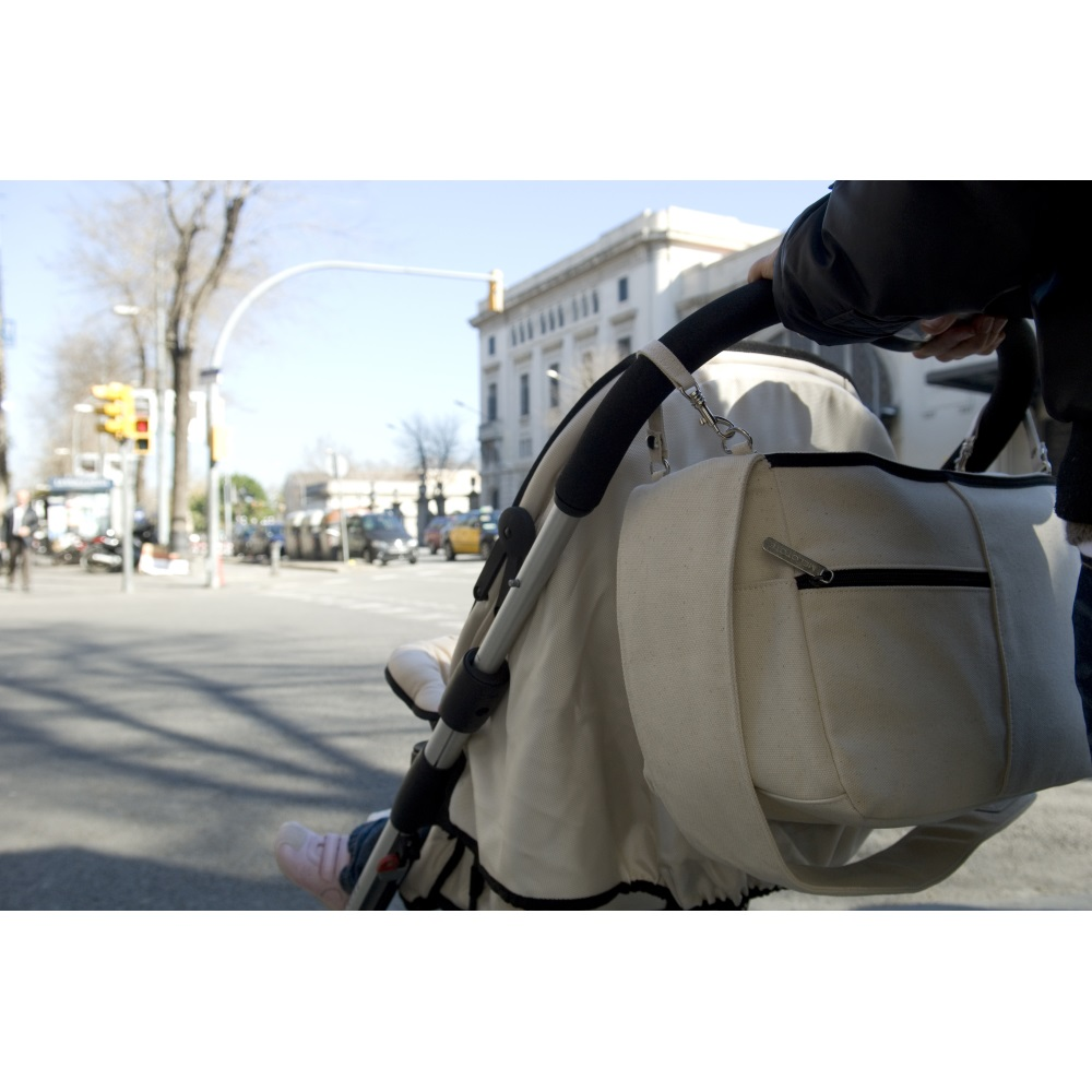 Ein Kinderwagen wird über die Straße geschoben. Eine beige Tasche hängt am Griff. Ein Kinderfuß mit rosa Schuh ist sichtbar.