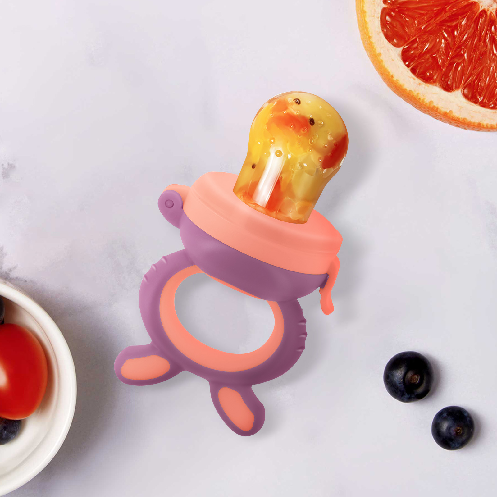 Un alimentatore per bambini con frutta arancione, mezzo pompelmo, mirtilli e una ciotola con pomodoro e mirtilli su una superficie bianca.