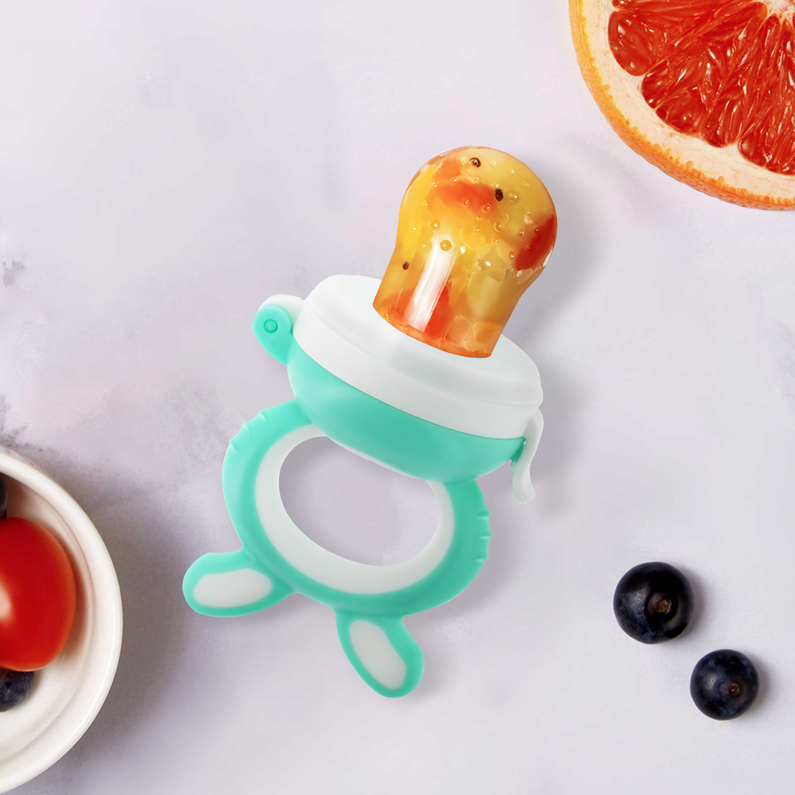Un alimentatore per neonati con frutta all'interno, una fetta di pompelmo, mirtilli e un pomodoro in una ciotola bianca sono su una superficie bianca.