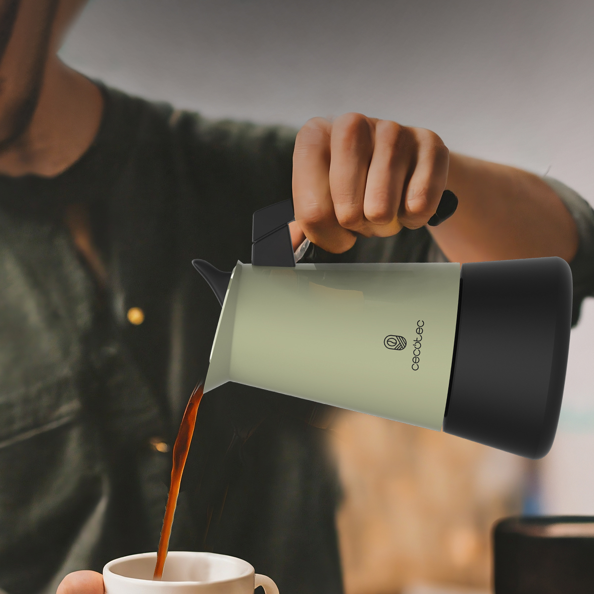 Una persona versa il caffè da una caffettiera verde e nera in una tazza bianca. La persona indossa una camicia verde.