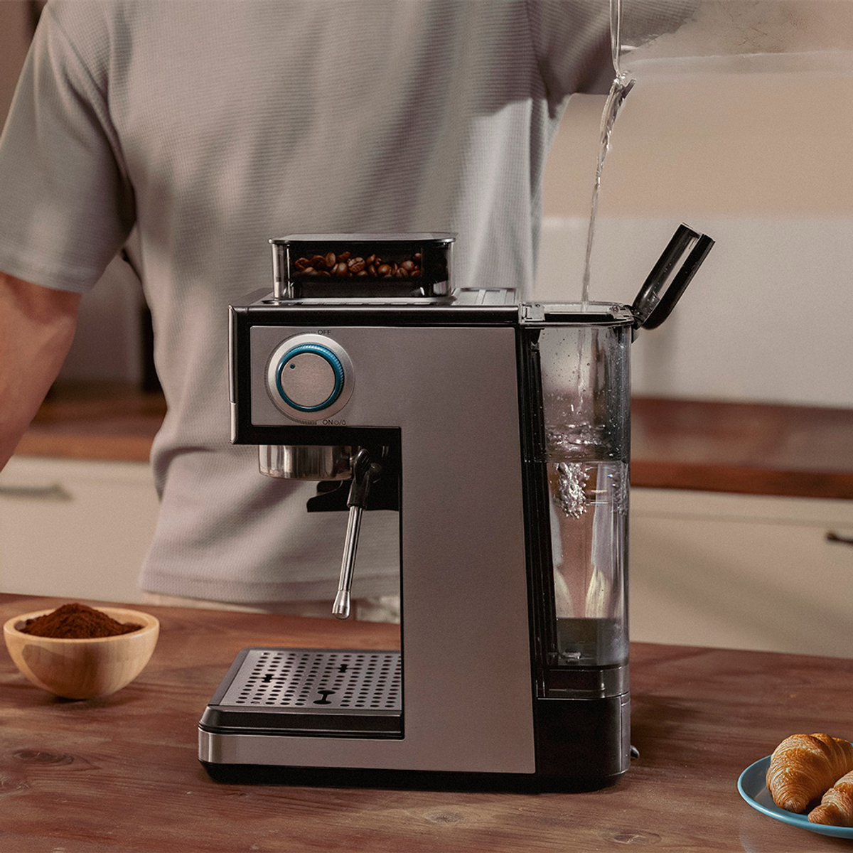 Una persona vierte agua en una máquina de espresso. El café molido está en un cuenco de madera. Cruasanes en un plato están en el fondo.