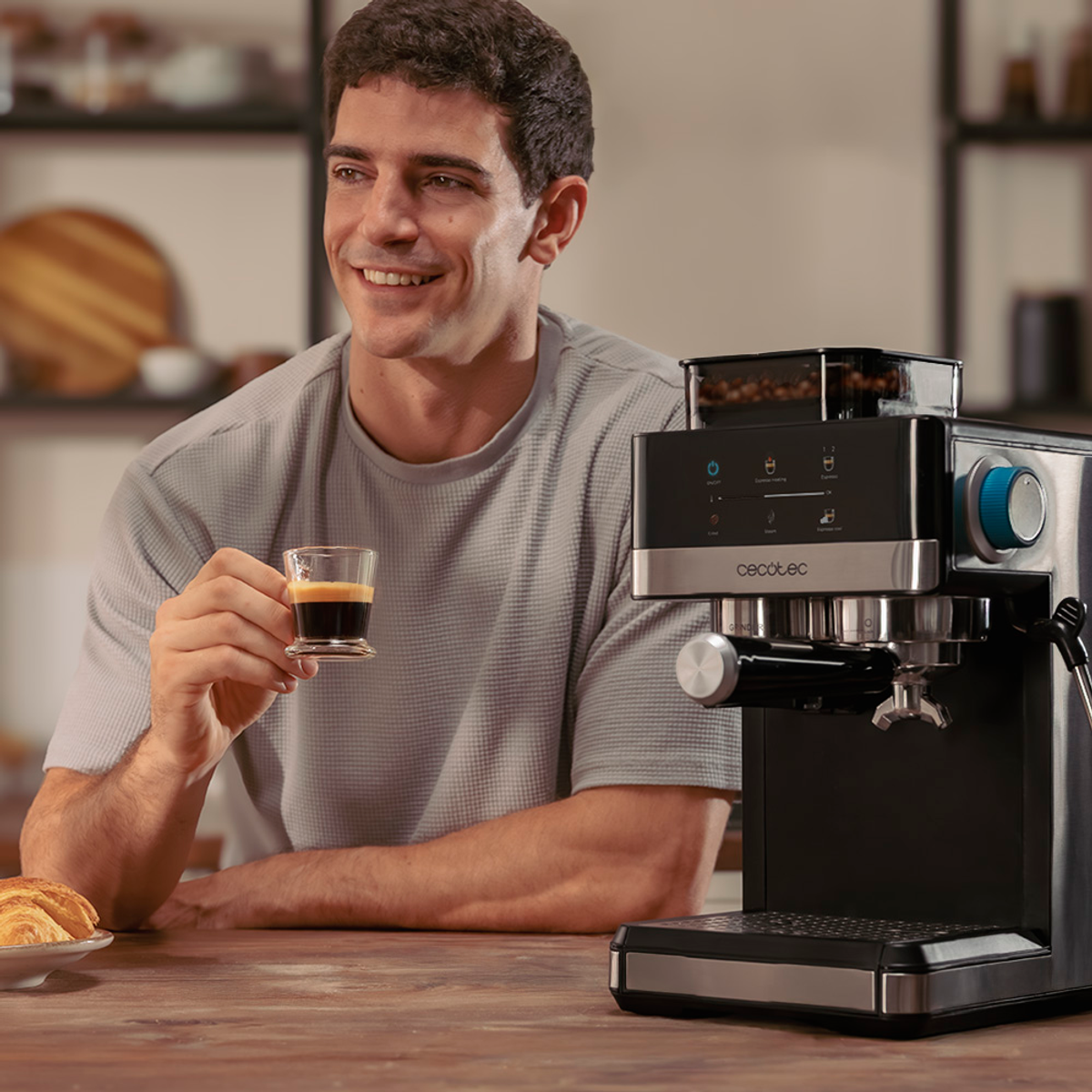 Un hombre sonríe, sosteniendo un espresso. Hay una cafetera a la derecha. Un cruasán a la izquierda.