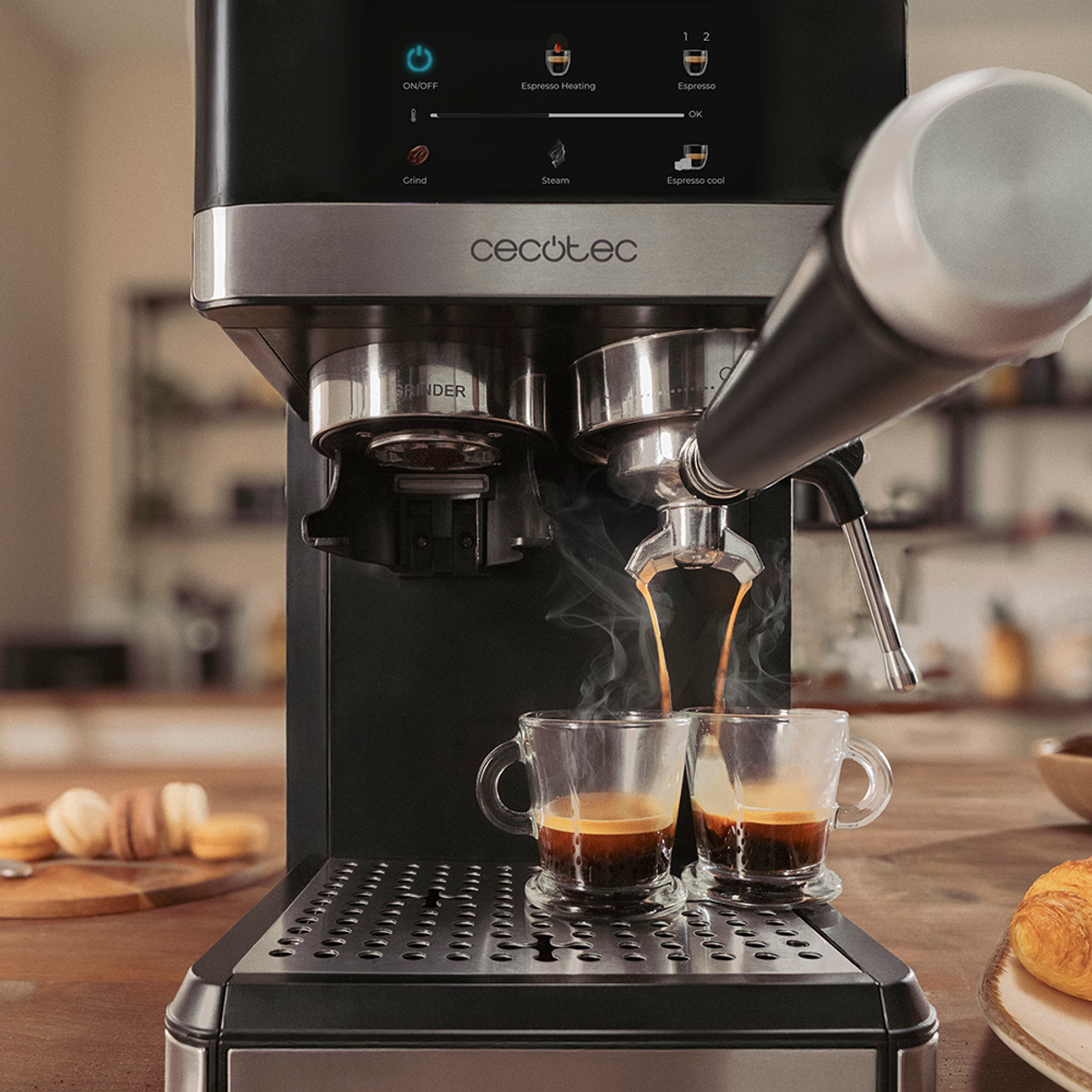 Una cafetera dispensando espresso en dos tazas de vidrio. Sale vapor. Un cruasán y galletas en la mesa.
