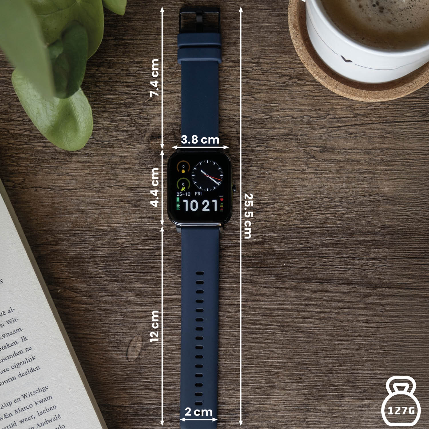 Een smartwatch met afmetingen op een houten oppervlak. Een kop koffie en een boek op de achtergrond. Het horloge toont de datum en tijd.