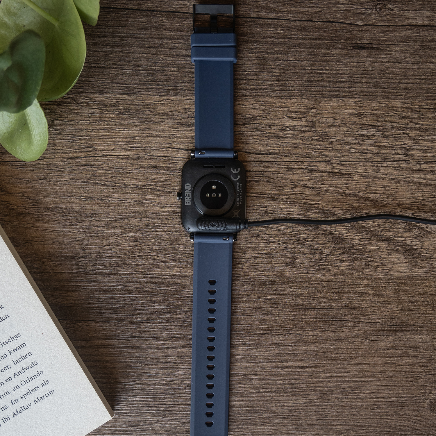 Een blauw smartwatch ligt op een houten oppervlak. Het is verbonden met een oplader. Een boek en groene bladeren liggen ernaast.