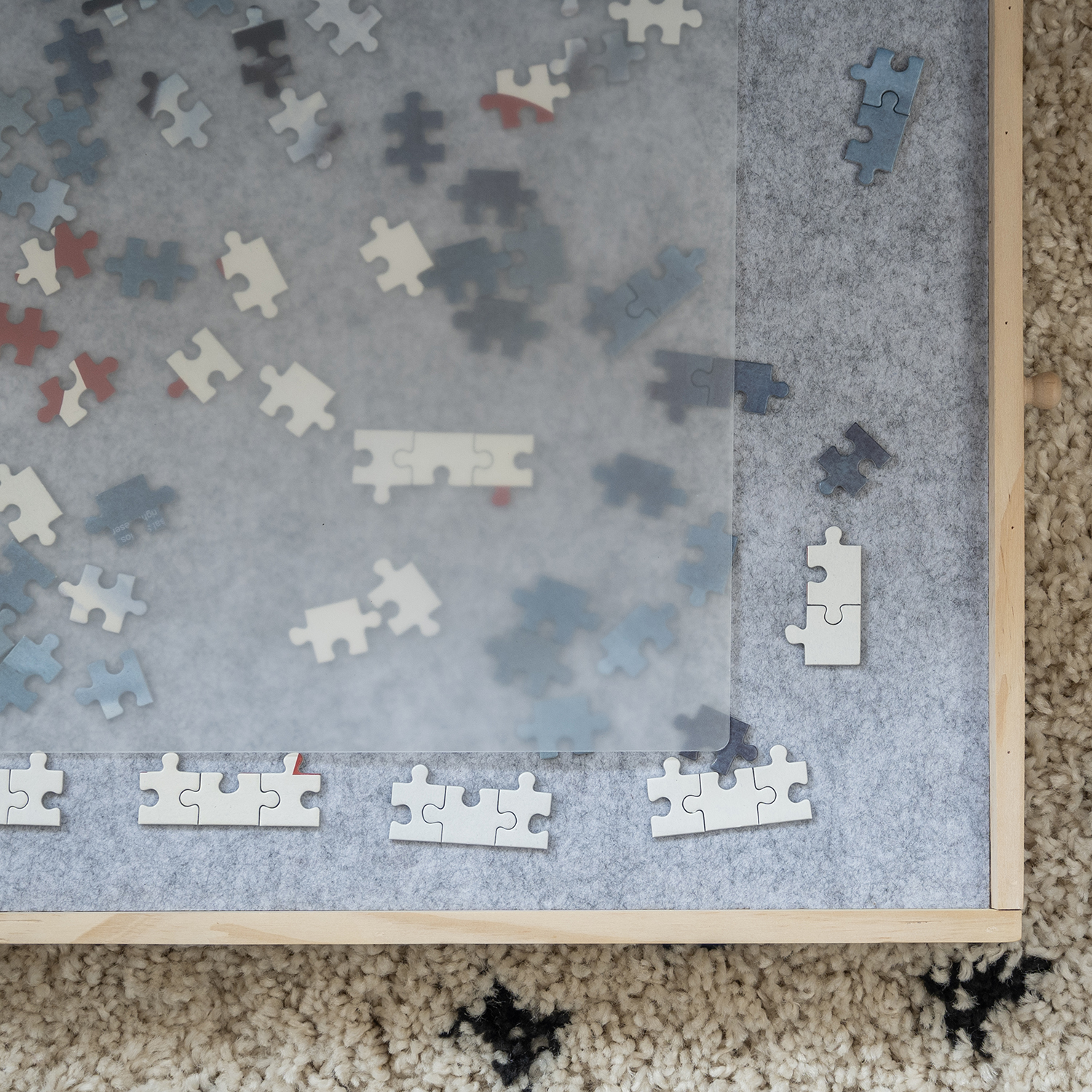 A partially completed puzzle on a gray felt board within a wooden frame. Scattered puzzle pieces are visible. The background is a patterned rug.