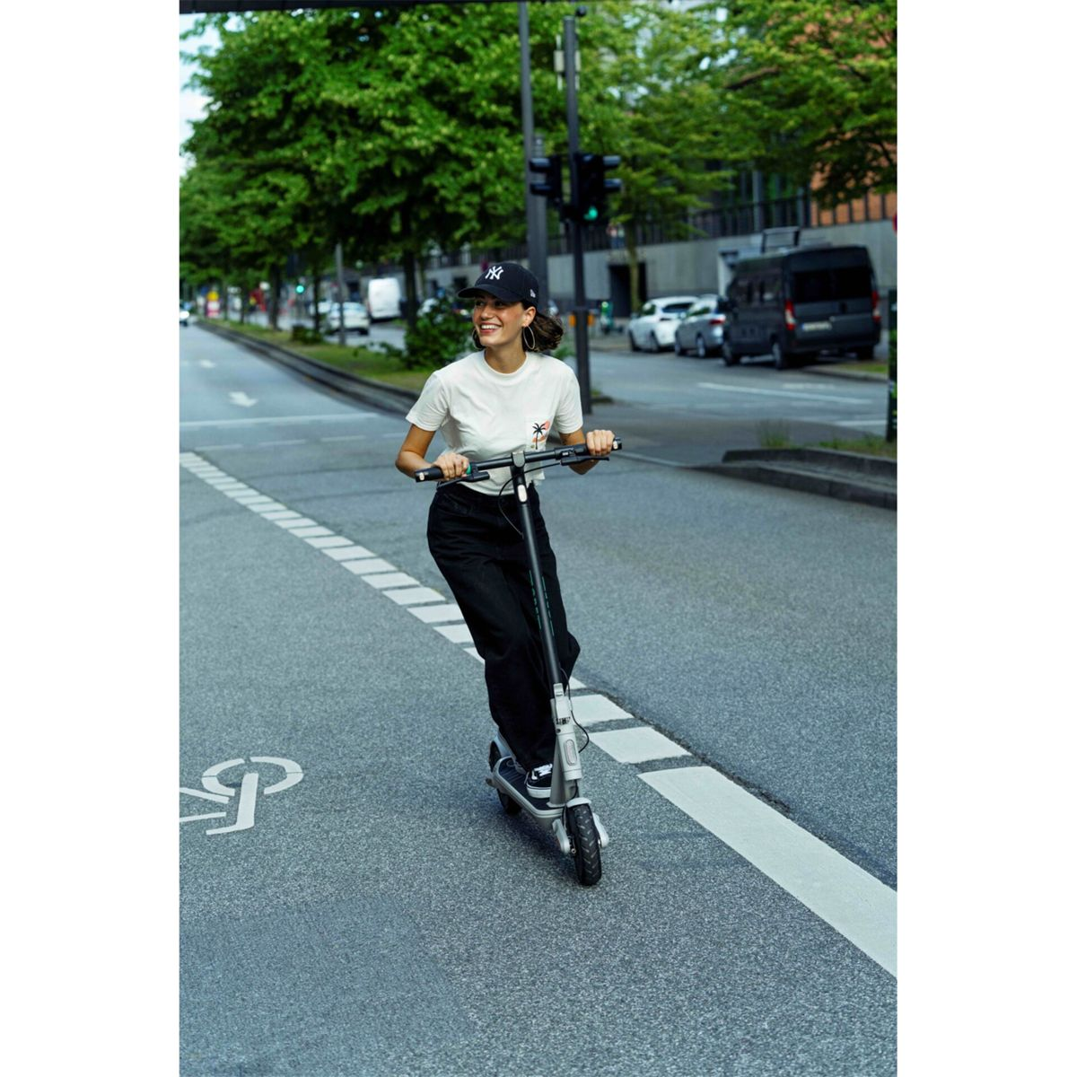 Eine Frau auf einem Roller lächelt, während sie auf einer Straße fährt. Sie trägt eine Kappe und schwarze Hosen. Bäume und Autos sind im Hintergrund.