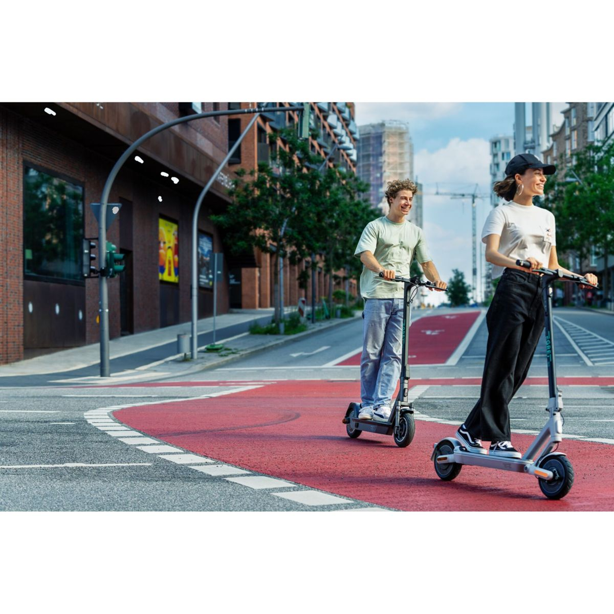 Zwei Personen fahren mit Elektrorollern auf einem roten Zebrastreifen in einer Stadt. Gebäude und Bäume säumen die Straße. Der Himmel ist blau.