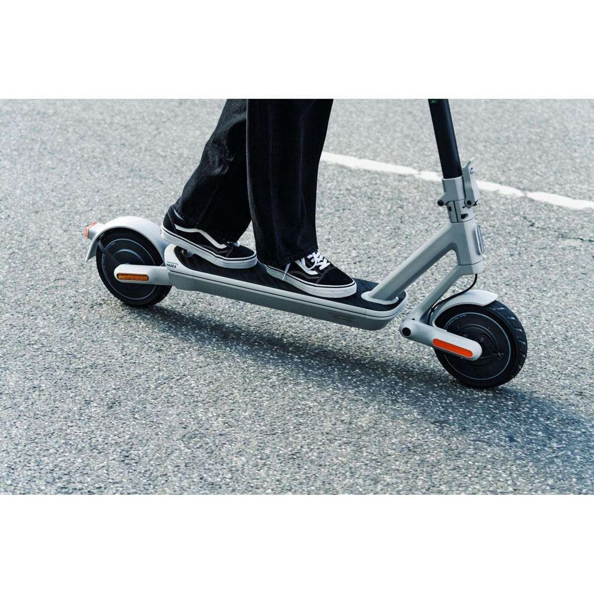 Person auf einem grauen Elektroroller mit schwarzen Rädern, trägt schwarze Hose und Vans-Schuhe, auf Asphalt.