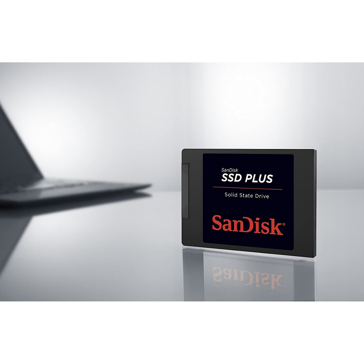 Czarny dysk SanDisk SSD Plus jest w centrum uwagi. Laptop jest rozmyty w tle. Dysk znajduje się na odblaskowej powierzchni.