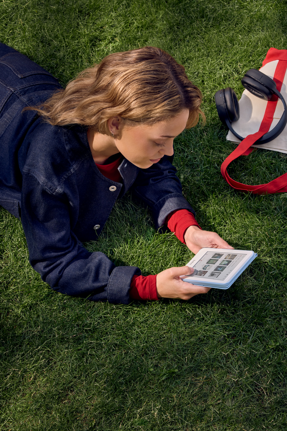 Kobieta leży na trawie, czytając czytnik e-booków. Ma na sobie dżinsową kurtkę i czerwoną koszulę. W pobliżu znajdują się słuchawki i torba.