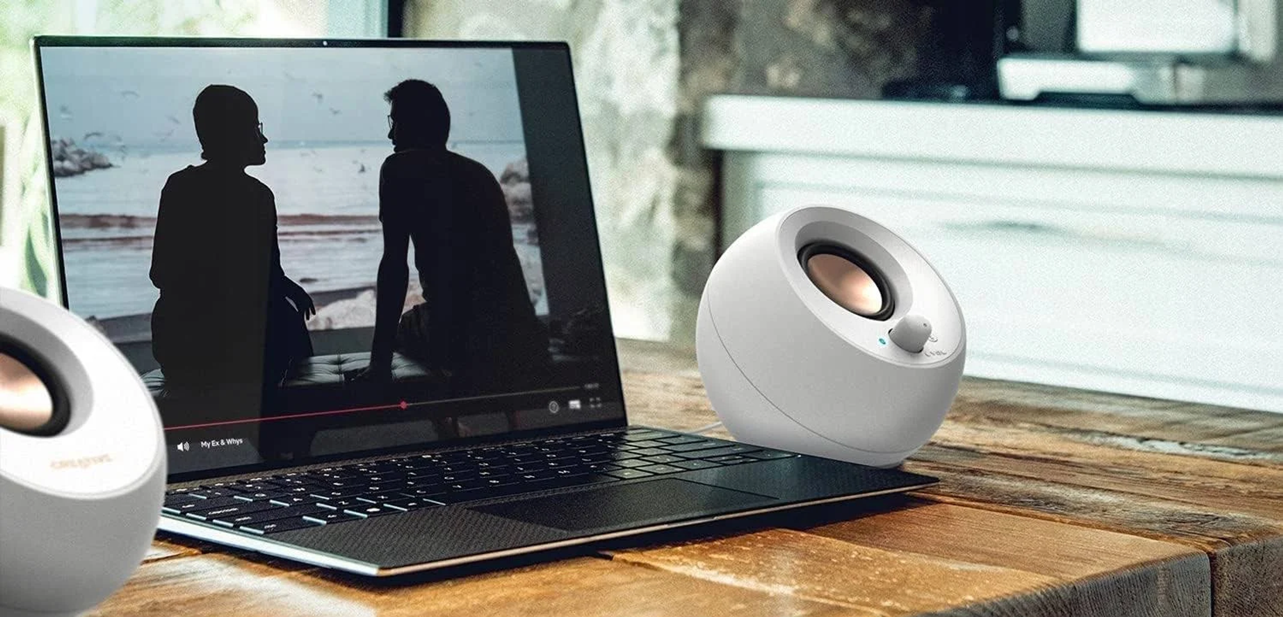 Laptop wyświetla scenę filmową. Dwa białe głośniki na drewnianym stole. Sylwetki ludzi na ekranie.