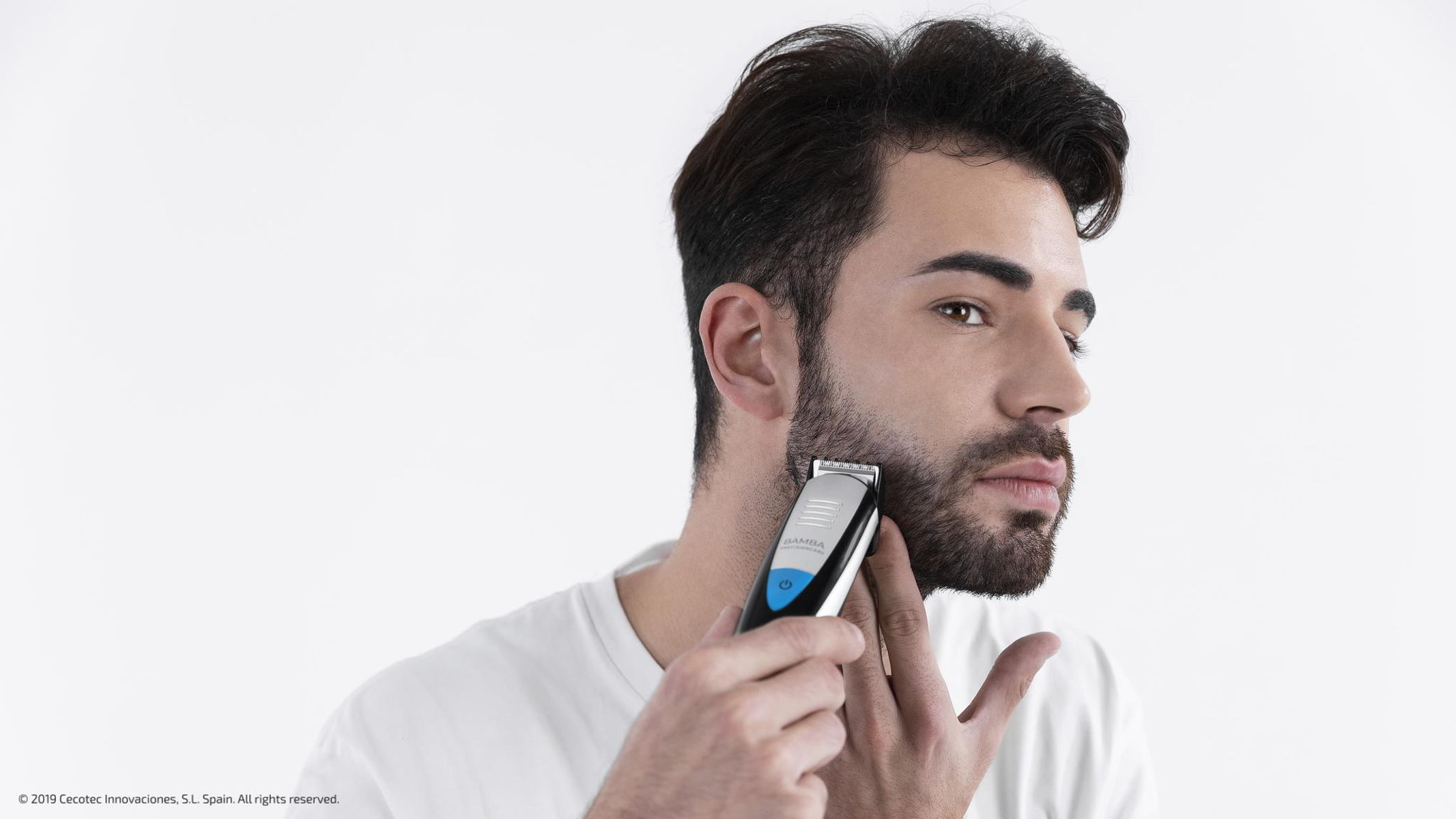 Un hombre se afeita la barba con una recortadora eléctrica. Sostiene la recortadora cerca de su mejilla. Fondo blanco.