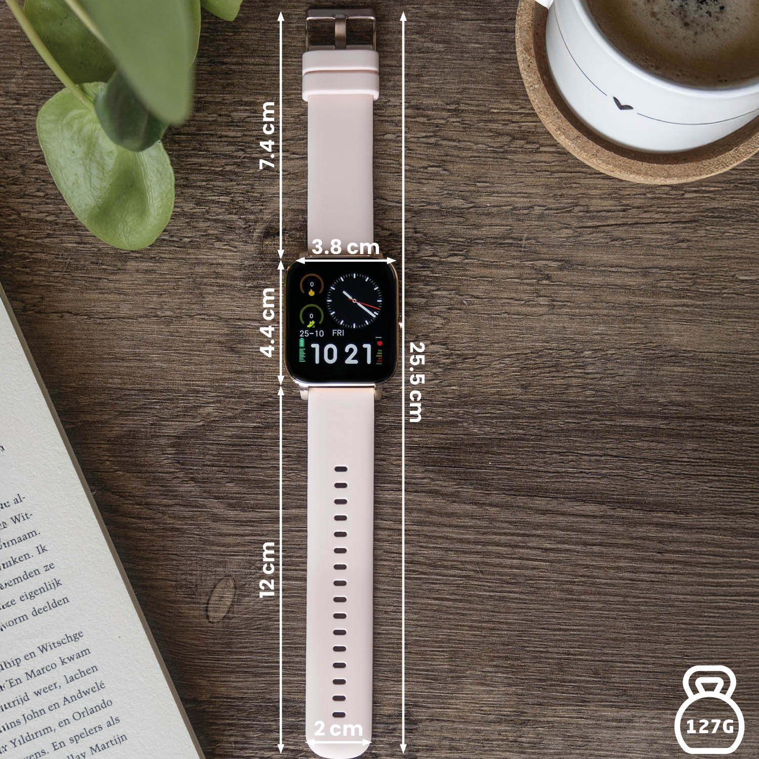 Een roze smartwatch ligt op een houten oppervlak. Afmetingen worden aangegeven. Een kopje koffie en een boek zijn ook aanwezig.