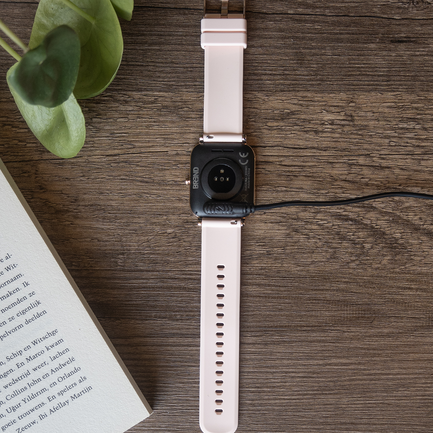 Een roze smartwatch met een zwarte oplader ligt op een houten tafel. Een boek en een groene plant zijn ook zichtbaar.