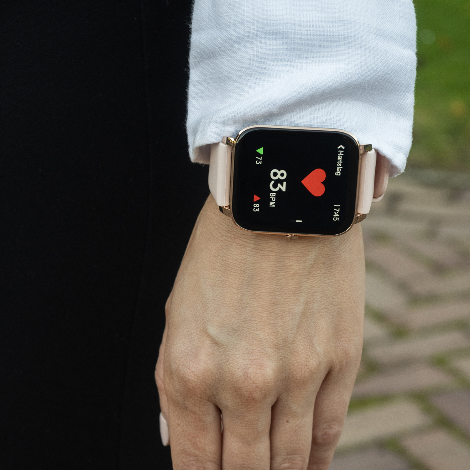 Een persoon draagt een smartwatch met hartslagmeting. Het horloge is goudkleurig met een roze bandje. De achtergrond toont donkere kleding en groen.