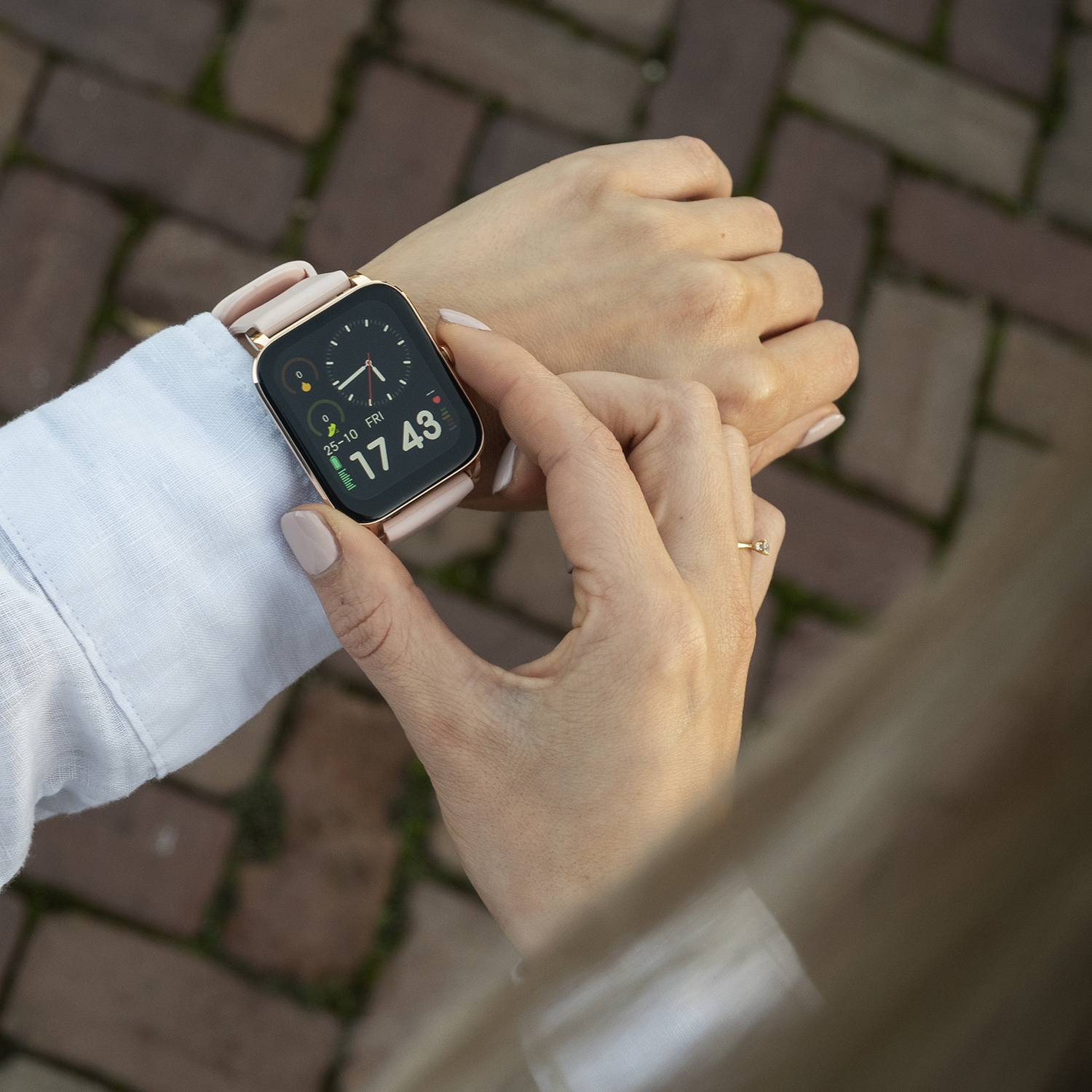 Pols van een persoon met een smartwatch die de tijd weergeeft. Het horloge heeft een roze bandje en de achtergrond is een bakstenen pad.