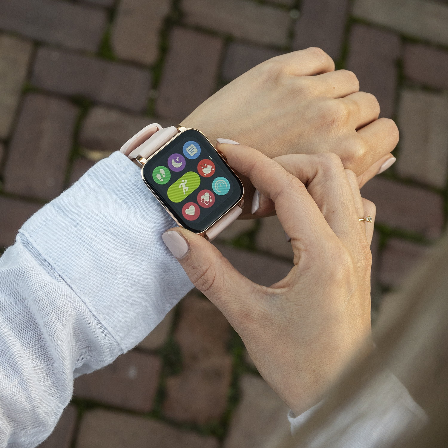 Een vrouw die een smartwatch met gezondheidspictogrammen draagt op een bakstenen pad, raakt het scherm aan. Ze draagt een lichtblauw overhemd.