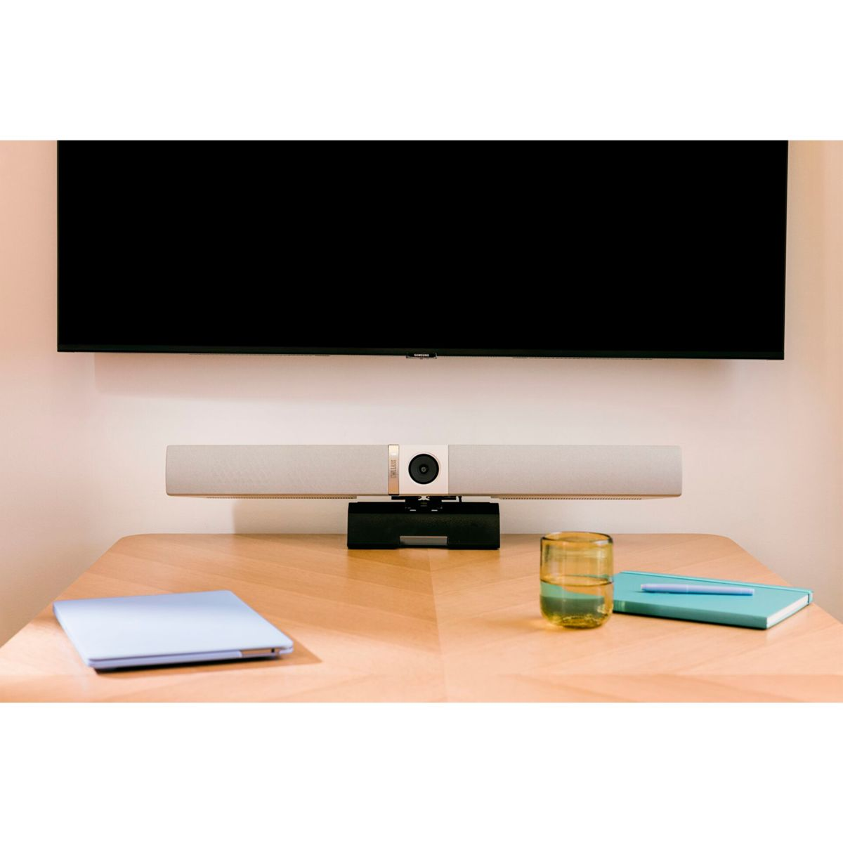 Ein Videokonferenz-Setup mit Bildschirm, Soundbar, Laptop, Wasserglas und Notizbuch auf einem Holztisch.