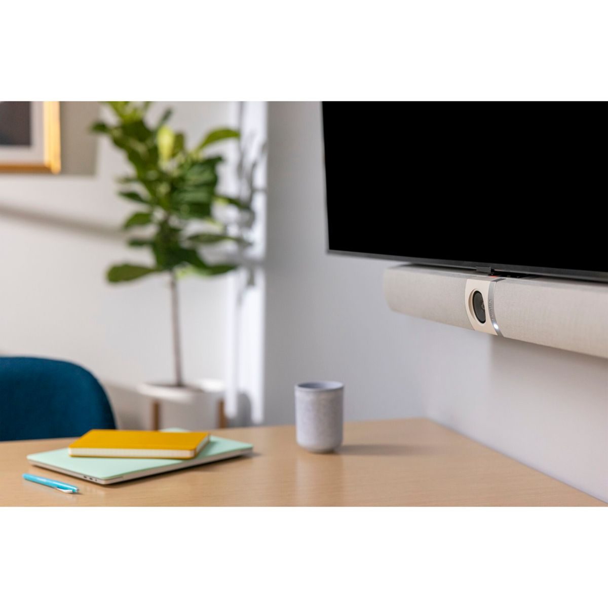 Ein Fernseher mit einer Soundbar, einem Notizbuch, einem Stift und einer Tasse auf einem Holztisch. Eine Pflanze in einem Topf ist im Hintergrund.