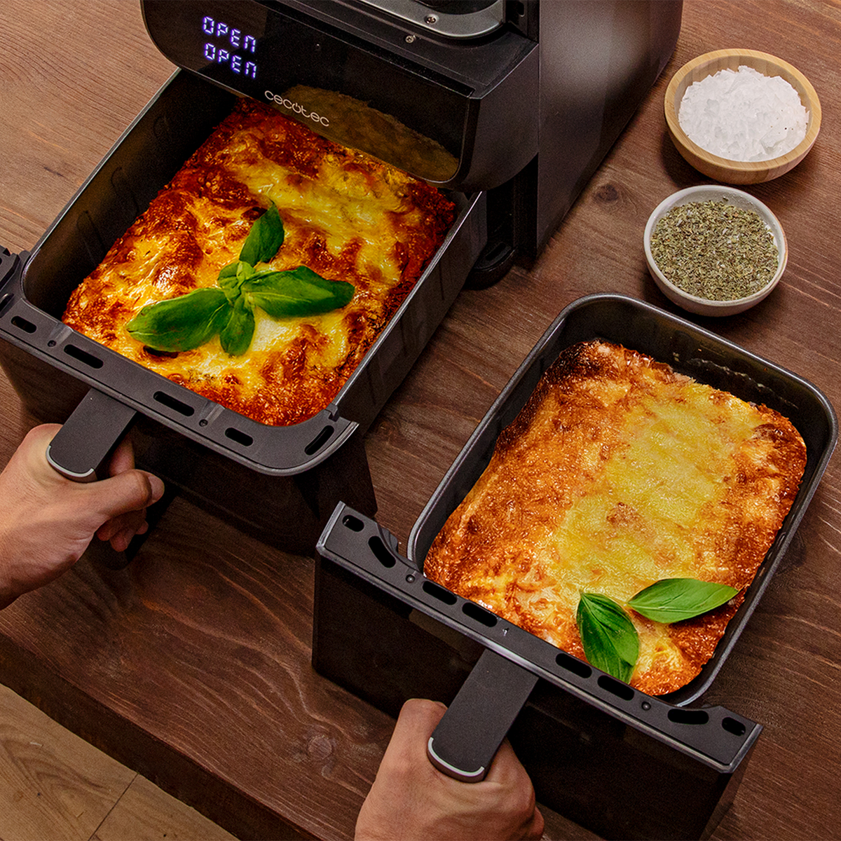 Due teglie di lasagne, una in una friggitrice ad aria aperta. Foglie di basilico in cima. Sale ed erbe aromatiche in ciotole su un tavolo di legno.