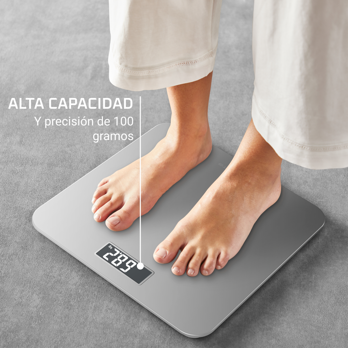 Persona in piedi su una bilancia digitale, viene visualizzato il peso. Bilancia grigia, pantaloni bianchi. Testo: Alta capacità e precisione di 100 grammi.