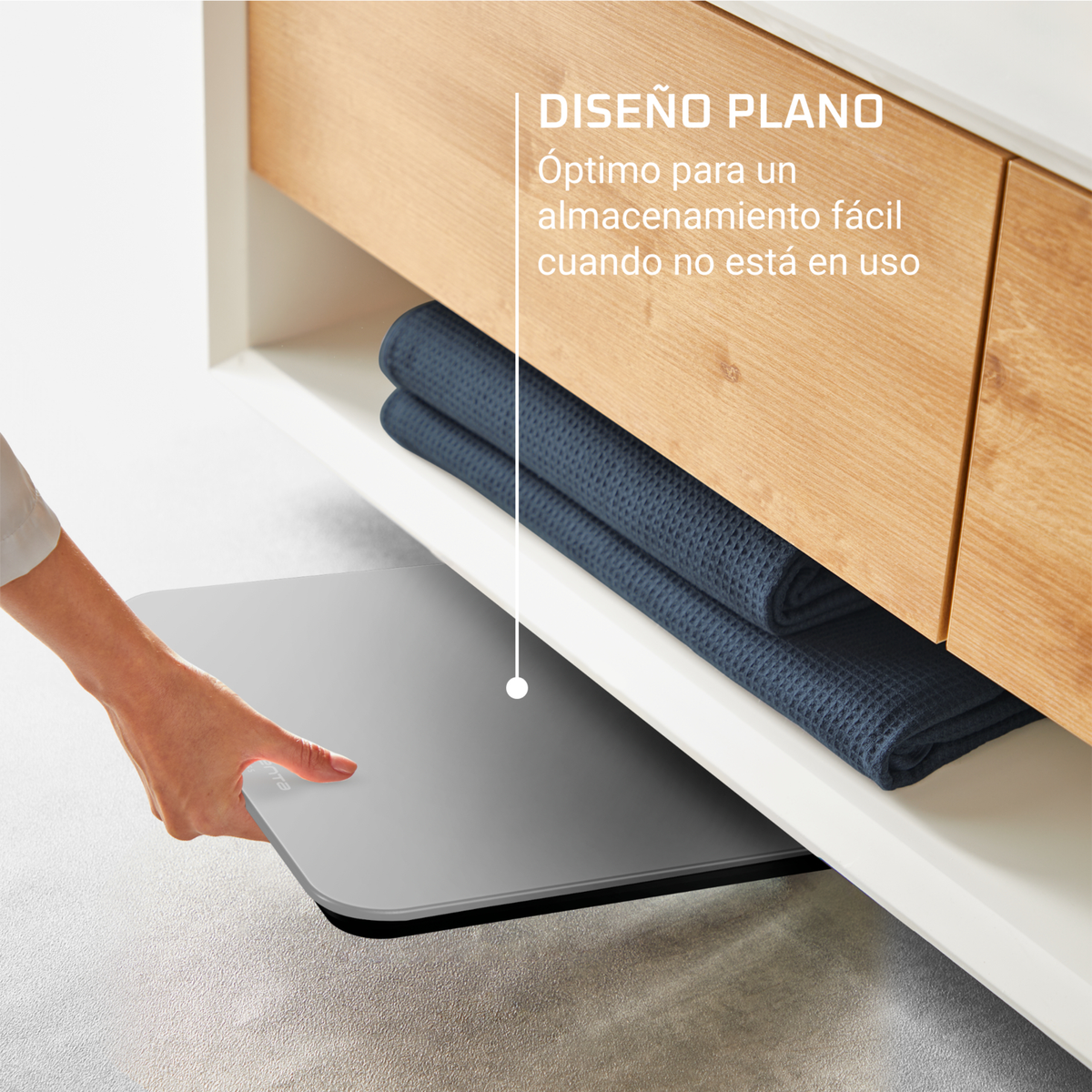 Una mano estrae una bilancia grigia da sotto un armadio con asciugamani. L'armadio è bianco con un piano in legno. È visibile anche del testo.