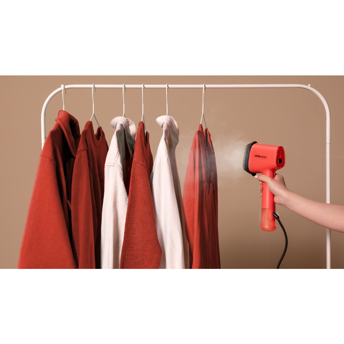 Una mano che tiene un vaporizzatore rosso stira i vestiti appesi a un appendiabiti. Diverse camicie rosse e bianche sono sugli appendini.
