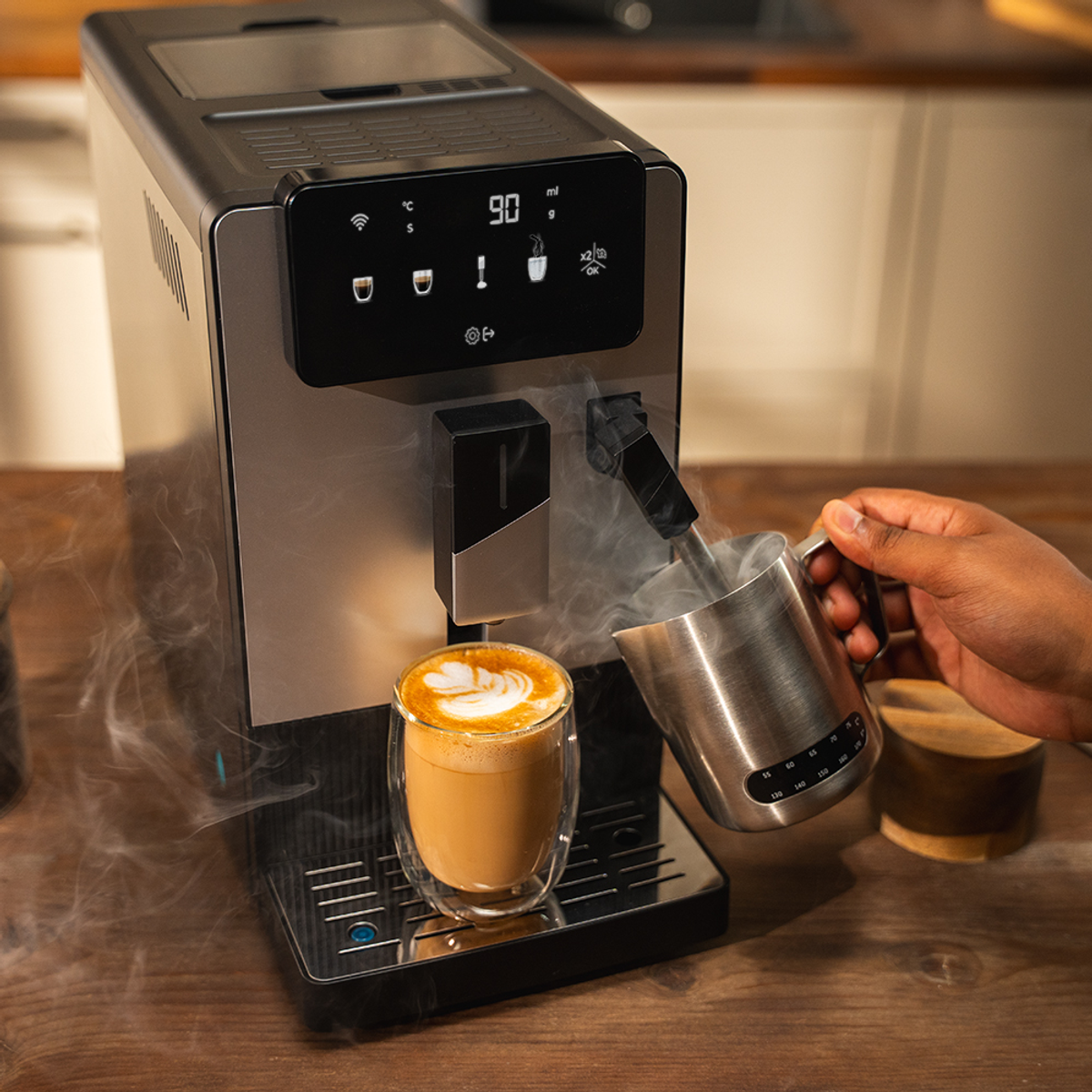 Una macchina da caffè che vaporizza latte in un bicchiere di latte macchiato. Una mano tiene un boccale di metallo. Lo sfondo è una cucina.