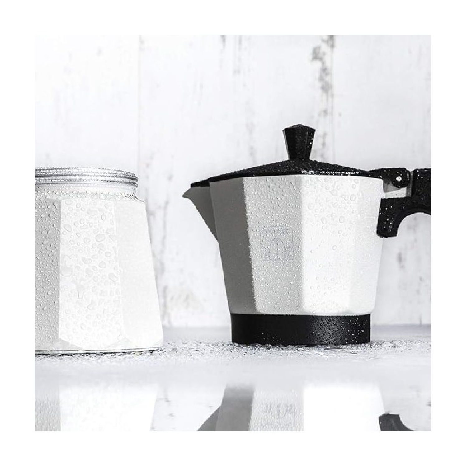 Macchina da caffè bianca e nera con goccioline d'acqua e un barattolo di vetro. Entrambi su una superficie riflettente, contro uno sfondo in legno chiaro.