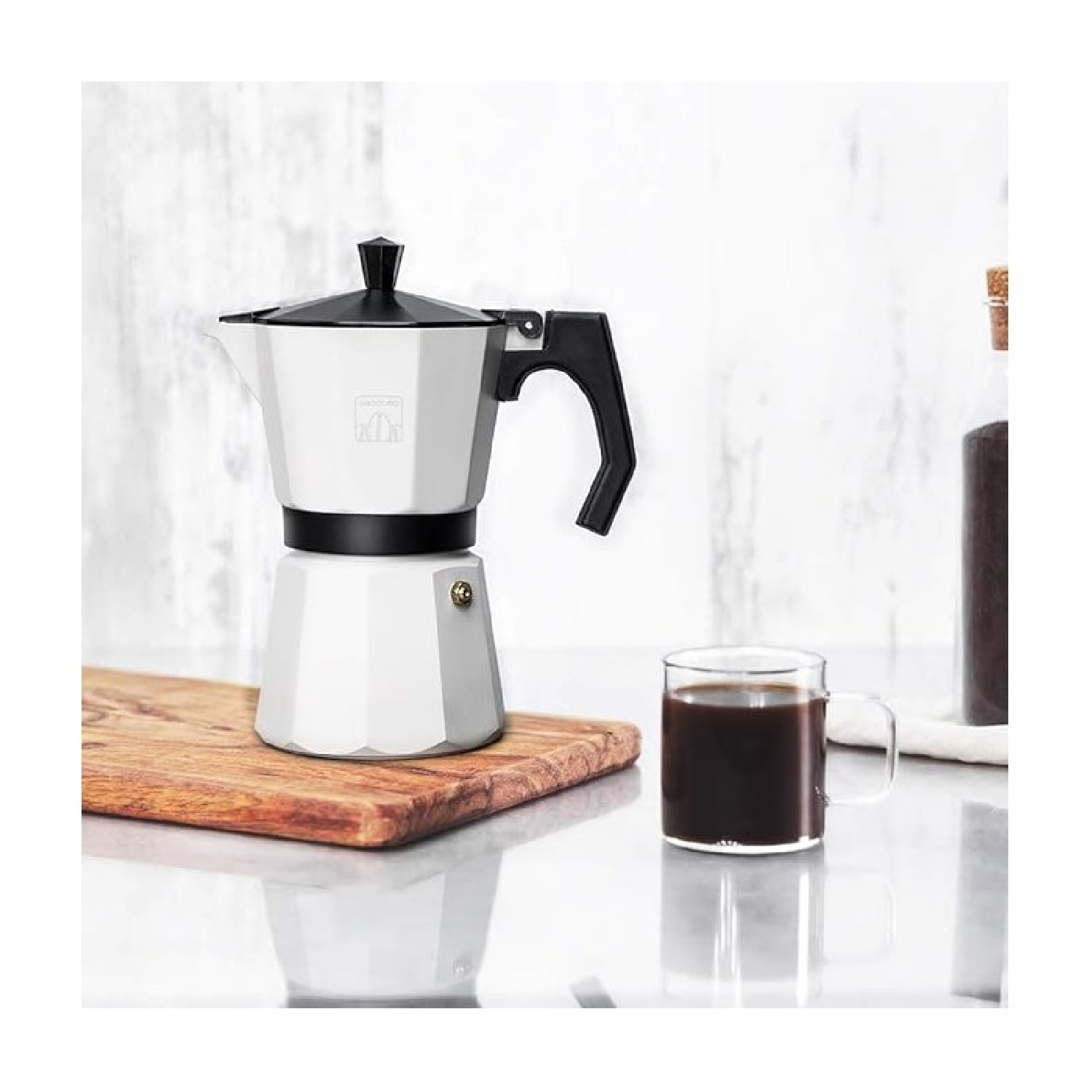 Macchina per caffè bianca su tagliere in legno, bicchiere di caffè e bottiglia su superficie bianca. Manico e coperchio neri.