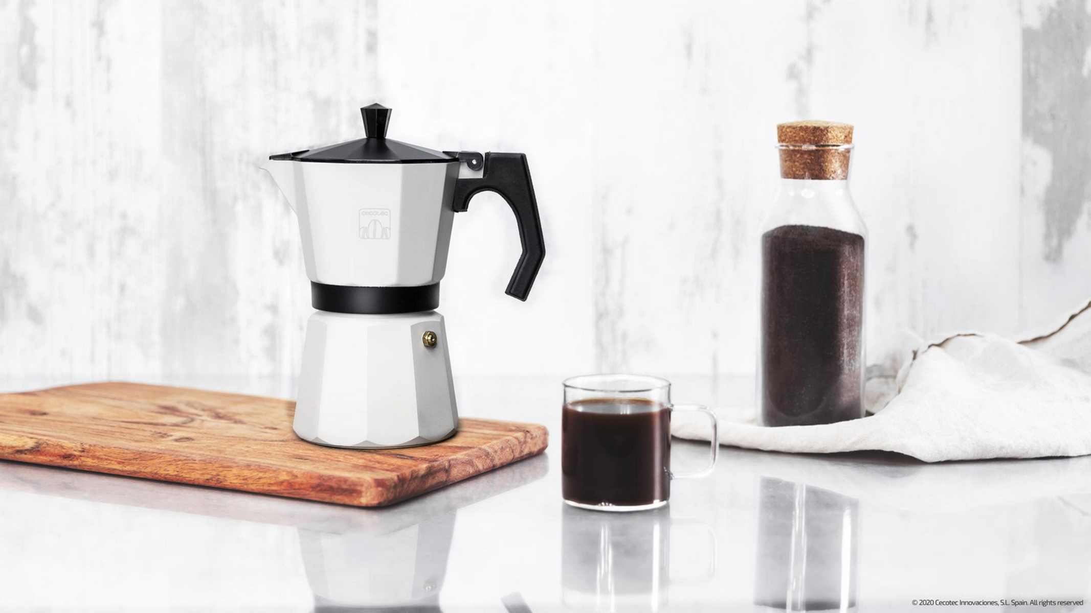 Una caffettiera bianca, una tazza di caffè, un barattolo di chicchi di caffè e un tagliere di legno. Lo sfondo è bianco.