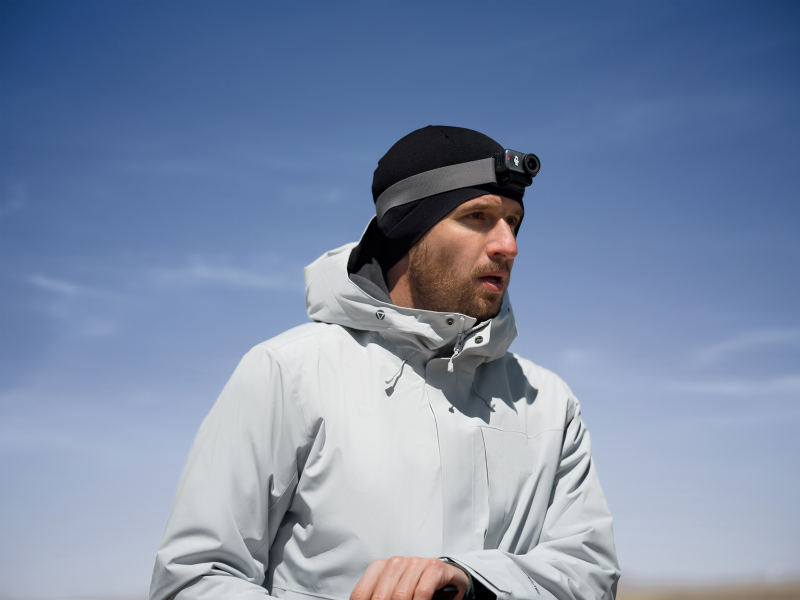 Mężczyzna w czarnej czapce z czołówką i szarej kurtce na tle błękitnego nieba. Ma brodę i patrzy w bok.