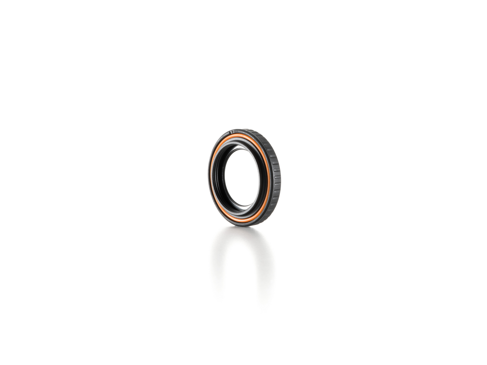 Un objeto circular negro y naranja, posiblemente un componente de lente, descansa sobre una superficie blanca. Proyecta una sombra suave.