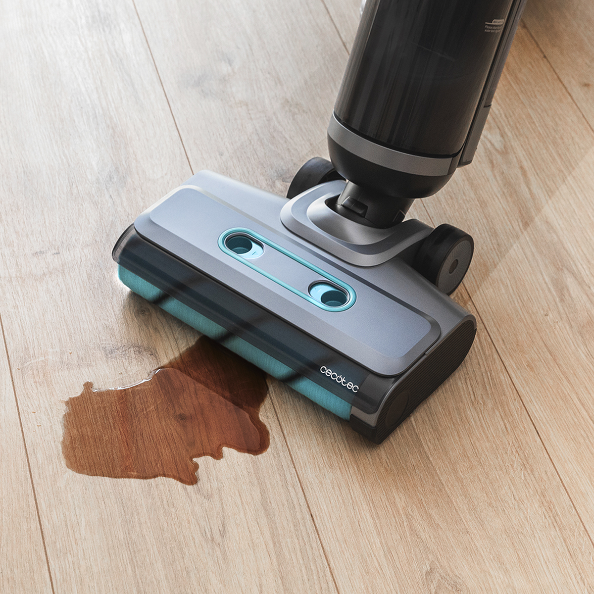 Una testina per aspirapolvere sta pulendo una macchia di liquido marrone su un pavimento in legno. L'aspirapolvere è grigio e nero.