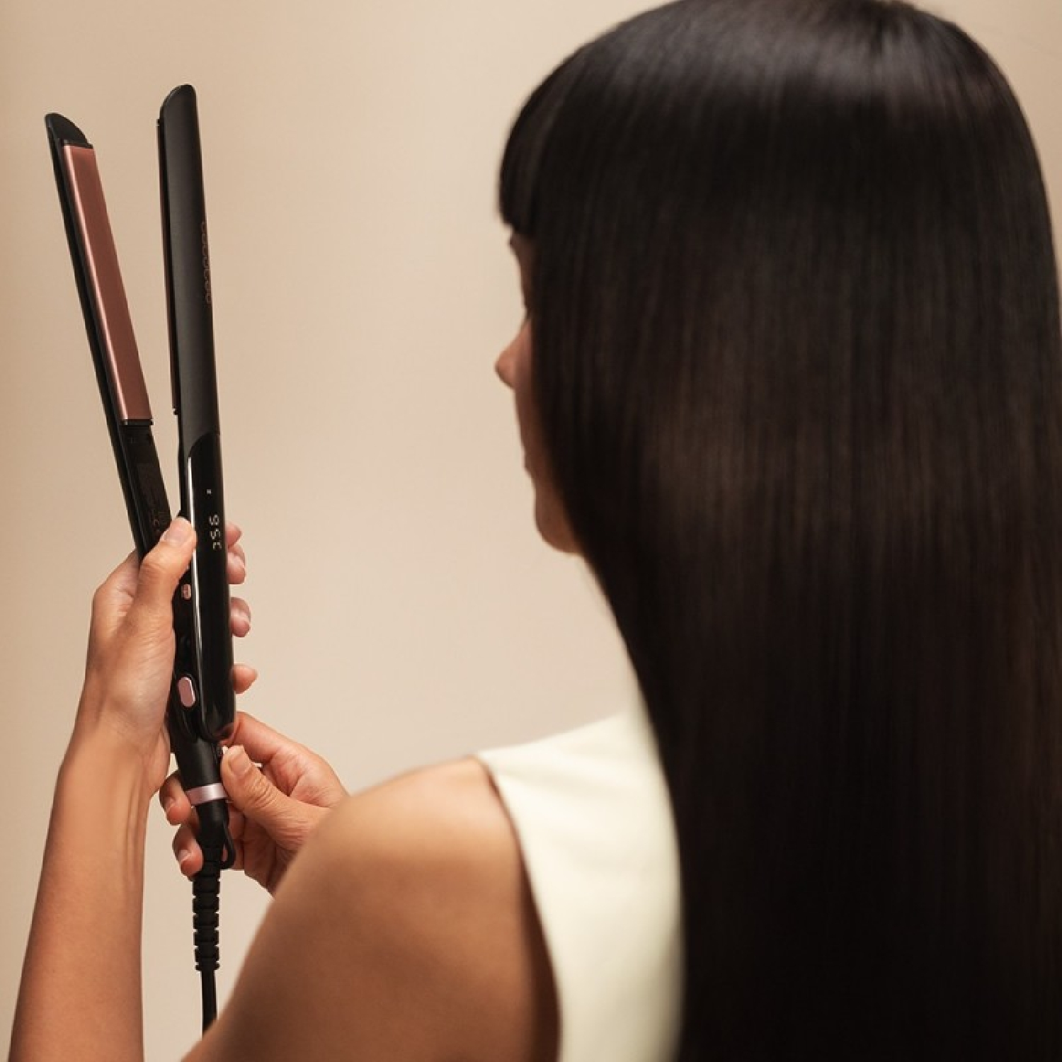 Una persona tiene in mano una piastra per capelli nera con piastre oro rosa. La persona ha capelli lunghi, lisci e neri.
