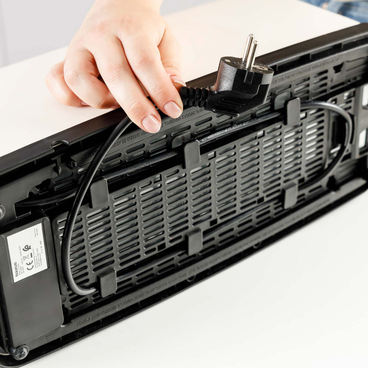 Una mano collega un cavo elettrico nero a un dispositivo. Il dispositivo ha un design a griglia e ventilazione nero. Sfondo bianco.