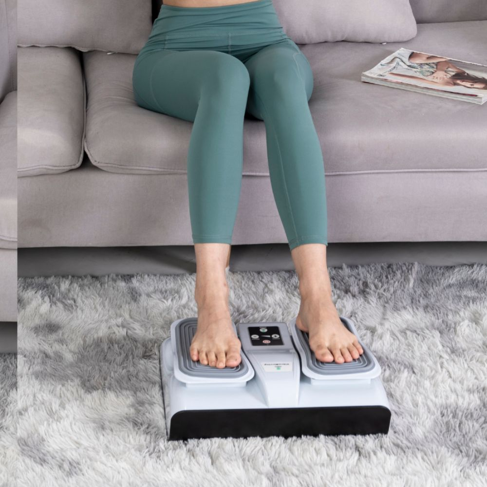 Una persona con leggings verde azulado en un sofá usa un masajeador de pies. Una revista está a su lado en un sofá gris.
