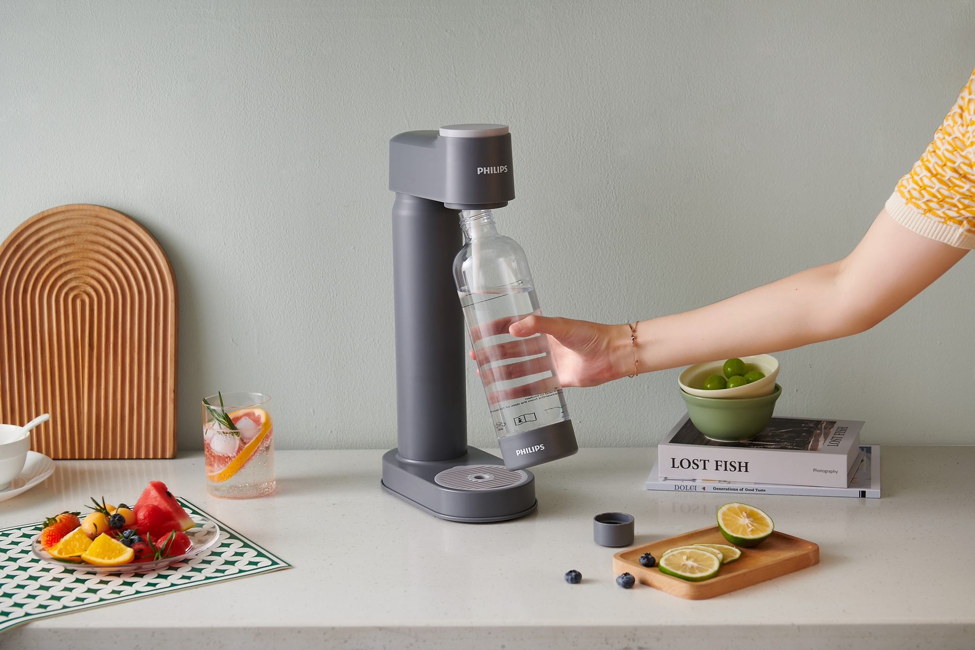 Una persona tiene in mano una bottiglia d'acqua sotto un produttore di soda Philips. Frutta e altri oggetti sono su un piano di lavoro bianco.