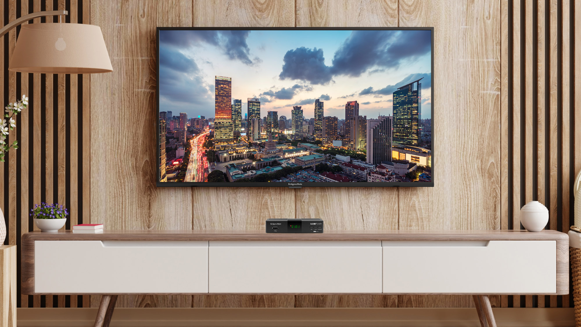 Telewizor wyświetla krajobraz miejski. Stoi na drewnianej szafce z lampą i dekoracjami. Ściana za nim jest również drewniana.